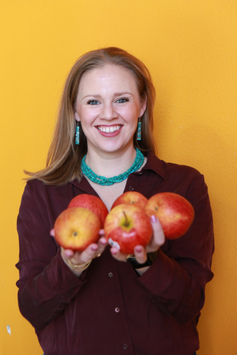 Kristen smiling and holding an assortment of fruit: watermelons, nectarines, lemons, and peppers